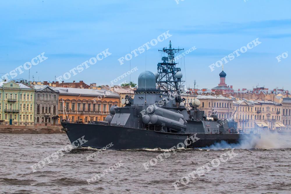 На фотографии изображен военный корабль, проходящий по реке Нева в Санкт-Петербурге. На фоне можно увидеть историческую застройку набережной с характерной архитектурой. Корабль участвует в демонстрации, подчеркивая связь города с военно-морской историей России. Нева, являясь главной водной артерией Петербурга, нередко становится местом проведения торжественных парадов флота.